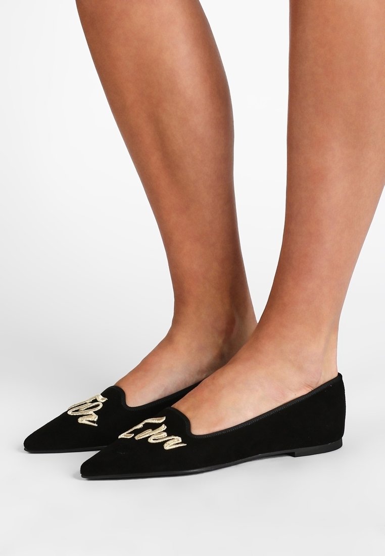 Chaussures plates en daim noir avec un bout pointu et un design découpé. Présentent des lettres brodées en or sur la tige. Semelle plate et lisse.