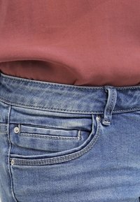 Primer plano de la cintura y el área del bolsillo de unos pantalones vaqueros azules con un lazo, combinados con una camisa de color rojo-rosado metida por dentro.