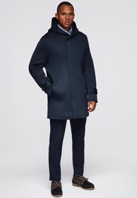 Abrigo con capucha de color azul marino con una textura suave, bolsillos frontales y cierres a presión, combinado con pantalones oscuros y zapatos marrones de cordones.