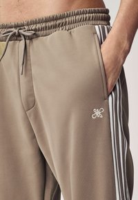 Taupe színű melegítőnadrág, puha anyagból készült, elasztikus derékkal, oldalsó zsebekkel és fehér csíkozással az oldalán.