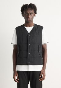 Vesta neagră matlasată cu nasturi tip snaps, având buzunare laterale, purtată peste un tricou alb cu dungi. Design simplu și material texturat.