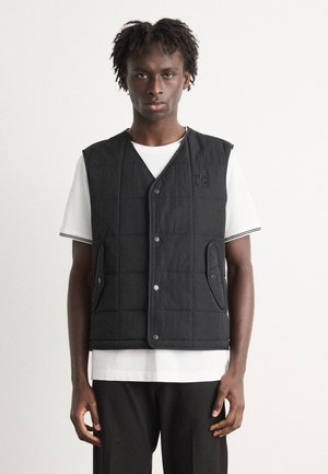 Gilet matelassé noir avec des boutons-pression, comportant des poches latérales, porté par-dessus un t-shirt rayé blanc. Design simple et tissu texturé.
