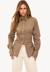 Brown striped button-up shirt with gathered detailing at the waist. Long sleeves and a collared neck. Worn with cream-colored pants.