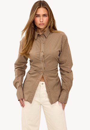 Brown striped button-up shirt with gathered detailing at the waist. Long sleeves and a collared neck. Worn with cream-colored pants.