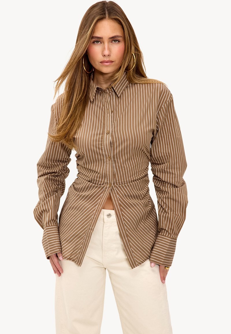 Brown striped button-up shirt with gathered detailing at the waist. Long sleeves and a collared neck. Worn with cream-colored pants.