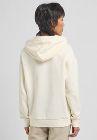 Felpa color crema con cappuccio arrotondato, maniche lunghe e polsini a costine. Il tessuto sembra morbido, con una vestibilità rilassata e un design minimalista.