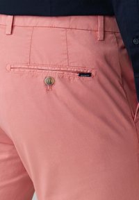 Primo piano della tasca posteriore di pantaloni rosa corallo con bottone e piccola etichetta nera Hackett, abbinati a una camicia blu navy scuro.