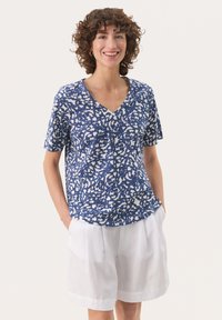 Blå och vit mönstrad kortärmad topp med v-ringning. Tyget har en texturerad look, parat med ljusa, löst sittande shorts.