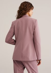 Prilagojen svetlo roza blazer s strukturiranimi rameni, enim zračnim razporkom na hrbtu in gladko teksturo, nošen s hlačami v istem odtenku.