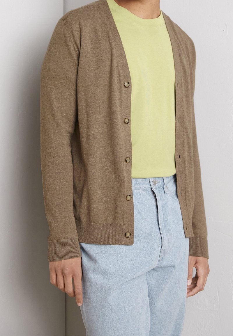 Hnědý cardigan s knoflíkovou přední částí, rukávy s žebrováním a měkkou texturou, nošený přes světle zelené tričko a v kombinaci se světle modrými džínami.