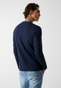 Granatowa koszula z długim rękawem i okrągłym dekoltem, o gładkiej teksturze i dopasowanym kroju, zestawiona z jasnoniebieskimi jeansami z detalem logo w kształcie trójkąta.