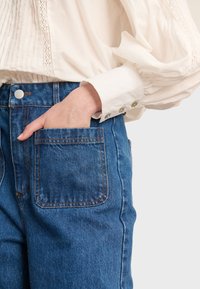 Tamnoplavih visokih traperica s vidljivim šavovima, prednjim džepom i metalnom kopčom, u kombinaciji s laganom teksturiranom bluzom.