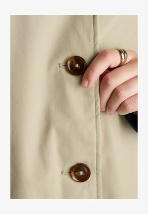 Hand mit einem goldenen Ring, der braune Knöpfe an beigem Stoff befestigt, Nahaufnahme der Vorderseite eines Mantels oder einer Jacke.