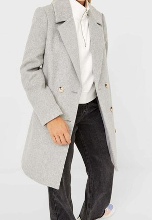 Personne portant un manteau en laine gris clair à double boutonnage, un haut blanc à col haut avec fermeture éclair, un jean noir à jambes larges et des chaussures blanches.