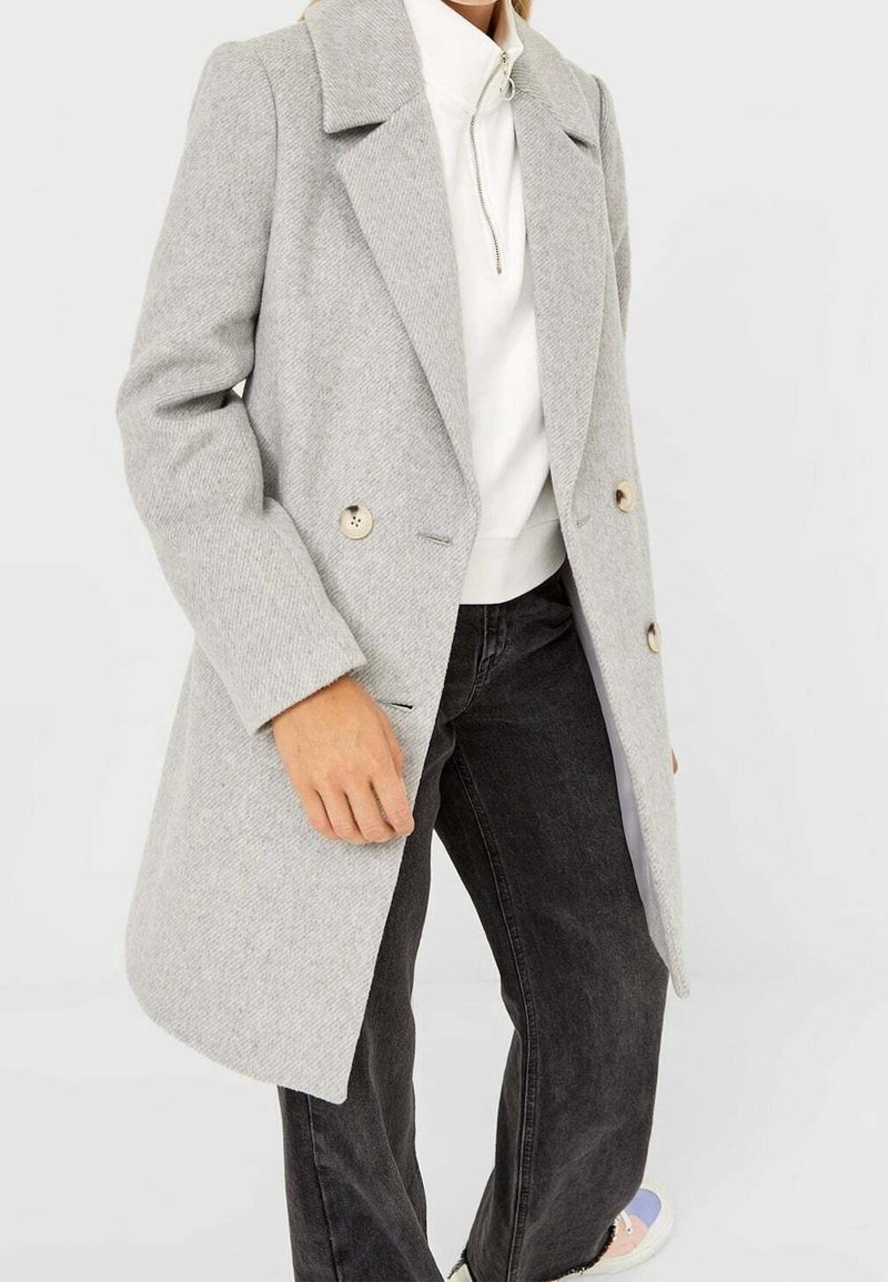 Personne portant un manteau en laine gris clair à double boutonnage, un haut blanc à col haut avec fermeture éclair, un jean noir à jambes larges et des chaussures blanches.