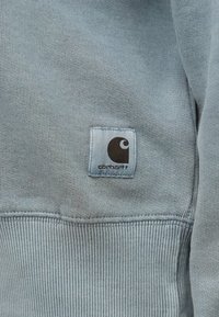 Tejido de sudadera gris con una textura suave; cuenta con una etiqueta cuadrada con un logotipo oscuro. Se pueden ver los puños y el dobladillo acanalados.