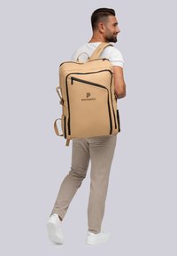 Mochila marrón con cremalleras y detalles negros, que cuenta con múltiples compartimentos y una correa ajustable, llevada por una persona con pantalones claros.