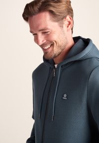 Dark teal zip-up hoodie made from soft fabric. Features a front zipper and drawstring hood, along with a small logo on the chest.