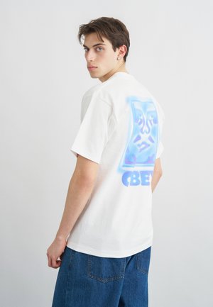 Νέος άνδρας με κοντά σκούρα μαλλιά, φοράει λευκό T-shirt με μπλε αφηρημένη εικόνα προσώπου και μπλε τζιν, γυρίζοντας να ρίξει μια ματιά πάνω από τον ώμο.