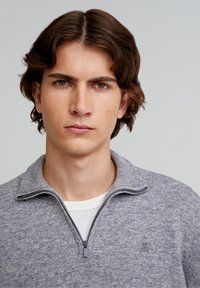 Grau zip-up Pullover aus weichem Stoff, mit hohem Kragen und einem kleinen Logodetail auf der Brust. Darunter ist ein weißes T-Shirt sichtbar.