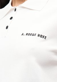 Λευκό πόλο πουκάμισο από μαλακό ύφασμα με κλασικό γιακά, διαθέτει μαύρα κουμπιά και κεντημένο κείμενο "A. ROEGE HOVE" σε μαύρο χρώμα.