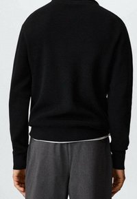 Pull noir côtelé à manches longues, col rond et coupe décontractée. Porté avec un pantalon gris, présentant un design simple et texturé.