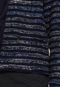 Veste bleu marine texturée avec des rayures horizontales blanches et bleues, comportant une petite poche zippée sur le devant.