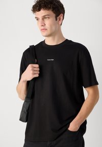 Black cotton t-shirt with short sleeves and a crew neckline. Features a white Calvin Klein logo on the chest. Held with a black strap.
