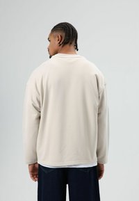 Beige oversized sweatshirt met een ronde hals, verlaagde schouders en lange mouwen, gemaakt van zachte stof, gedragen over donkere spijkerbroek.