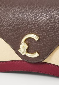 Sac à main en cuir avec des panneaux texturés marron et crème, présentant un accent en logo doré et un cordon bordeaux en bas.
