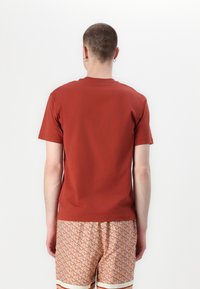 Tricou scurt de culoare ruginie, cu guler rotund, confecționat dintr-un material neted. Purtați-l cu pantaloni scurți cu modele geometrice în nuanțe de bej.