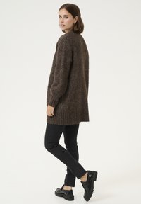 Hnědý pletený dlouhý cardigan s žebrovaným lemem a manžetami, texturovaná látka, v kombinaci s černými slim-fit džínami a robustními černými botami.