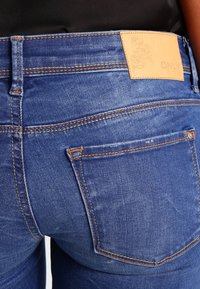 Jeans de denim azul oscuro con una cintura alta, que presentan costuras en contraste y un parche de cuero marrón con un diseño floral en relieve.