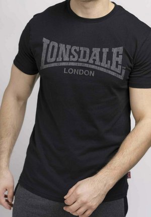 Mężczyzna ubrany w czarną koszulkę Lonsdale London i szare spodnie na jednolitym jasnym tle.
