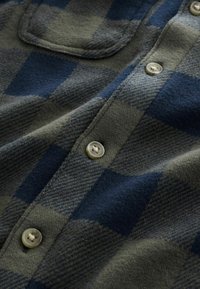 Flannel shirt in dark blue and olive green plaid. Features a chest pocket and beige buttons with a textured, soft fabric finish.