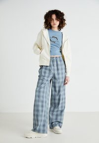 Licht crèmekleurige rits-hoodie, blauwe graphic T-shirt met dolfijnenprint, oversized geruite broek in blauw, groen en wit, gecombineerd met chunky witte sneakers.