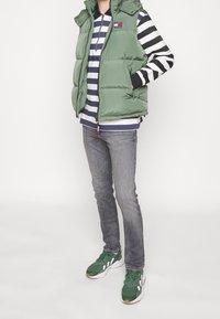 Osoba nosící zelenou nafouklou vestu, černo-bílé pruhované tričko s dlouhým rukávem, šedé džíny a zelené tenisky, stojící s rukama v kapsách.