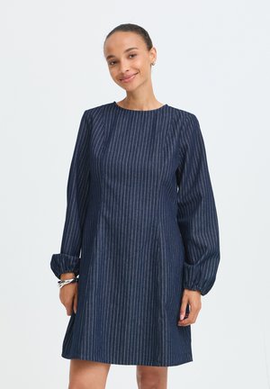 Femme souriante portant une robe bleu foncé à rayures fines, manches longues, poignets froncés et col rond, sur un fond uni.