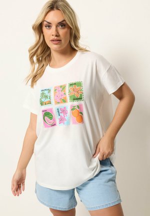 Blondynka w białej koszulce z kolorowymi tropikalnymi wzorami i jasnoniebieskich dżinsowych szortach na jednolitym tle.