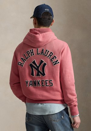 Różowa bluza z kapturem z czarnym nadrukiem "RALPH LAUREN" i "YANKEES" oraz dużym logo NY. Noszona z jasną koszulą dżinsową i ciemnymi dżinsami.