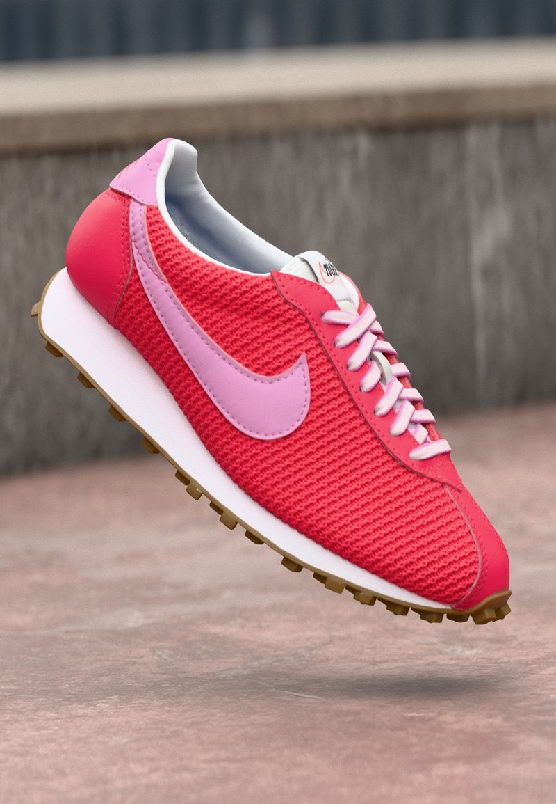 Jasne ružové a červené pletené tenisky s svetlo ružovým logom a šnúrkami, bielou podrážkou a hnedým vzorom, vznášajúce sa nad betónovým povrchom.