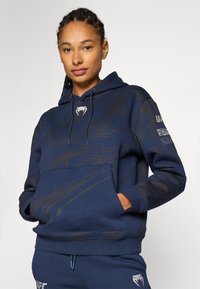 Felpa blu navy con un motivo testurizzato, tasca frontale e cordino regolabile. Presenta un logo sul petto e accenti stampati sulle maniche.