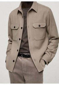 Beige wollen blazer met twee voorzakken, zwarte knopen en een puntige kraag, gecombineerd met een grijs shirt en corduroypantalon.