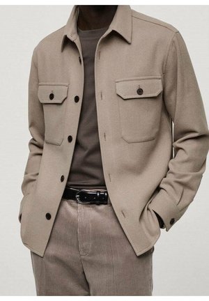 Beige wollen blazer met twee voorzakken, zwarte knopen en een puntige kraag, gecombineerd met een grijs shirt en corduroypantalon.