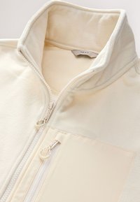 Fleecejacke in Cremefarbe mit hohem Kragen, durchgehendem Reißverschluss und einer Fronttasche, mit glatter Textur und schlichter Gestaltung.