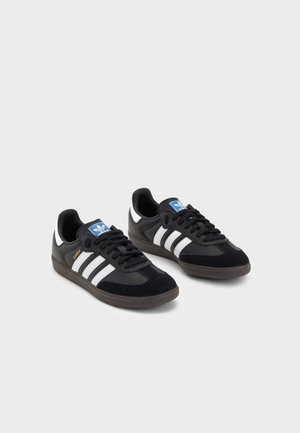 Et par sorte Adidas Samba sneakers med hvide striber, blåt logo på tungen og gummifarvede såler på en lys baggrund.