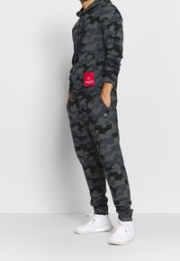 Haut à capuche et joggings camouflage en vert foncé et noir. Présente une poche avant et une étiquette rouge avec du texte blanc. Porté avec des baskets blanches.