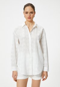 Camisa blanca de manga larga hecha de tela texturizada con un sutil patrón de hojas, que presenta un cierre de botones en la parte delantera y un cuello clásico.