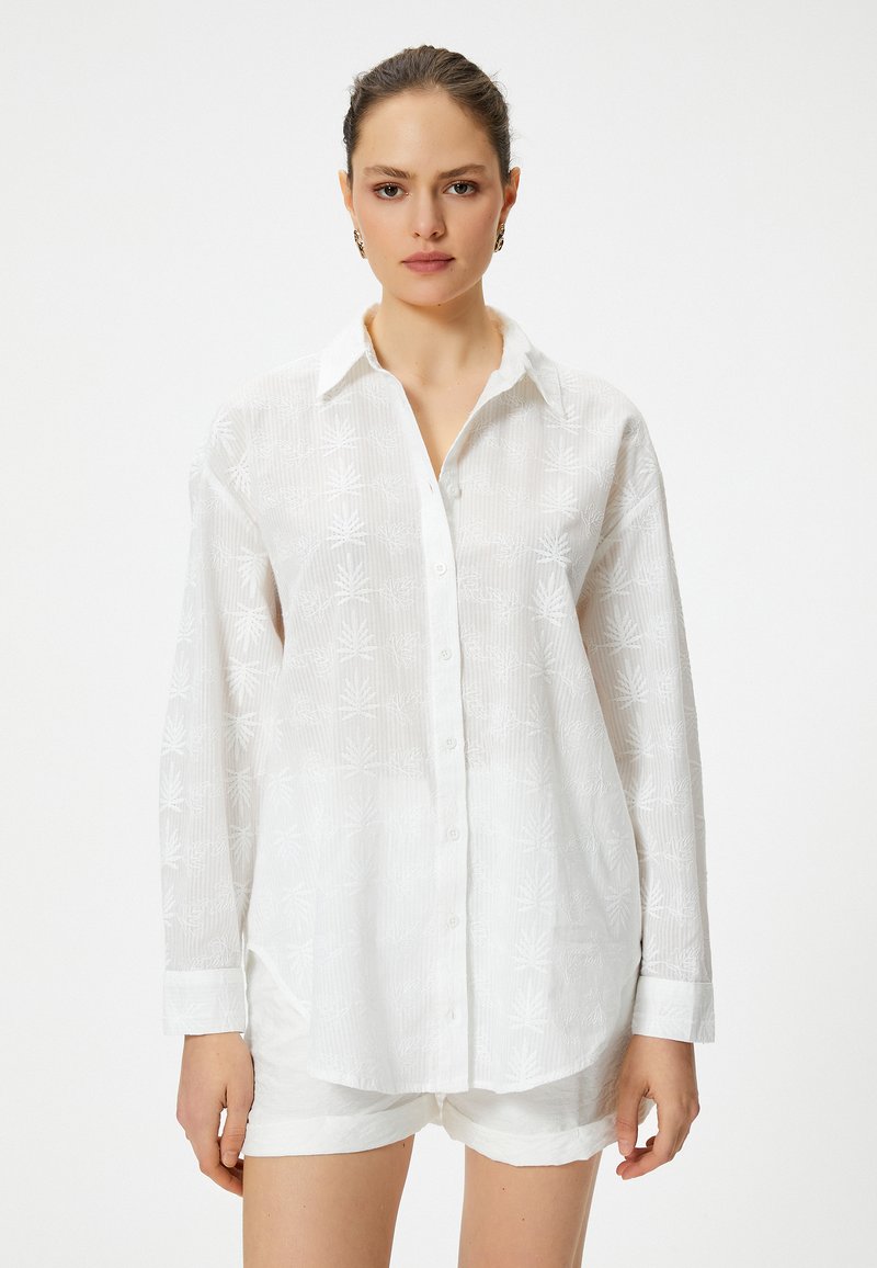 Camisa blanca de manga larga hecha de tela texturizada con un sutil patrón de hojas, que presenta un cierre de botones en la parte delantera y un cuello clásico.