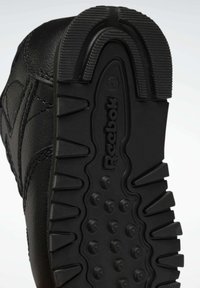 Svart gummisul med en texturerad mönster, med spår och cirkulära accenter, märkt med "Reebok"-logotypen.